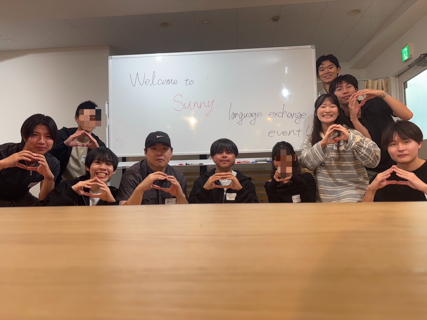 「Welcome to Sunny language exchange event!!」と書かれたホワイトボードの前で、参加者たちが手でハートを作って笑顔で写る集合写真 / Group photo of participants making heart shapes with their hands in front of a whiteboard reading “Welcome to Sunny language exchange event!!”